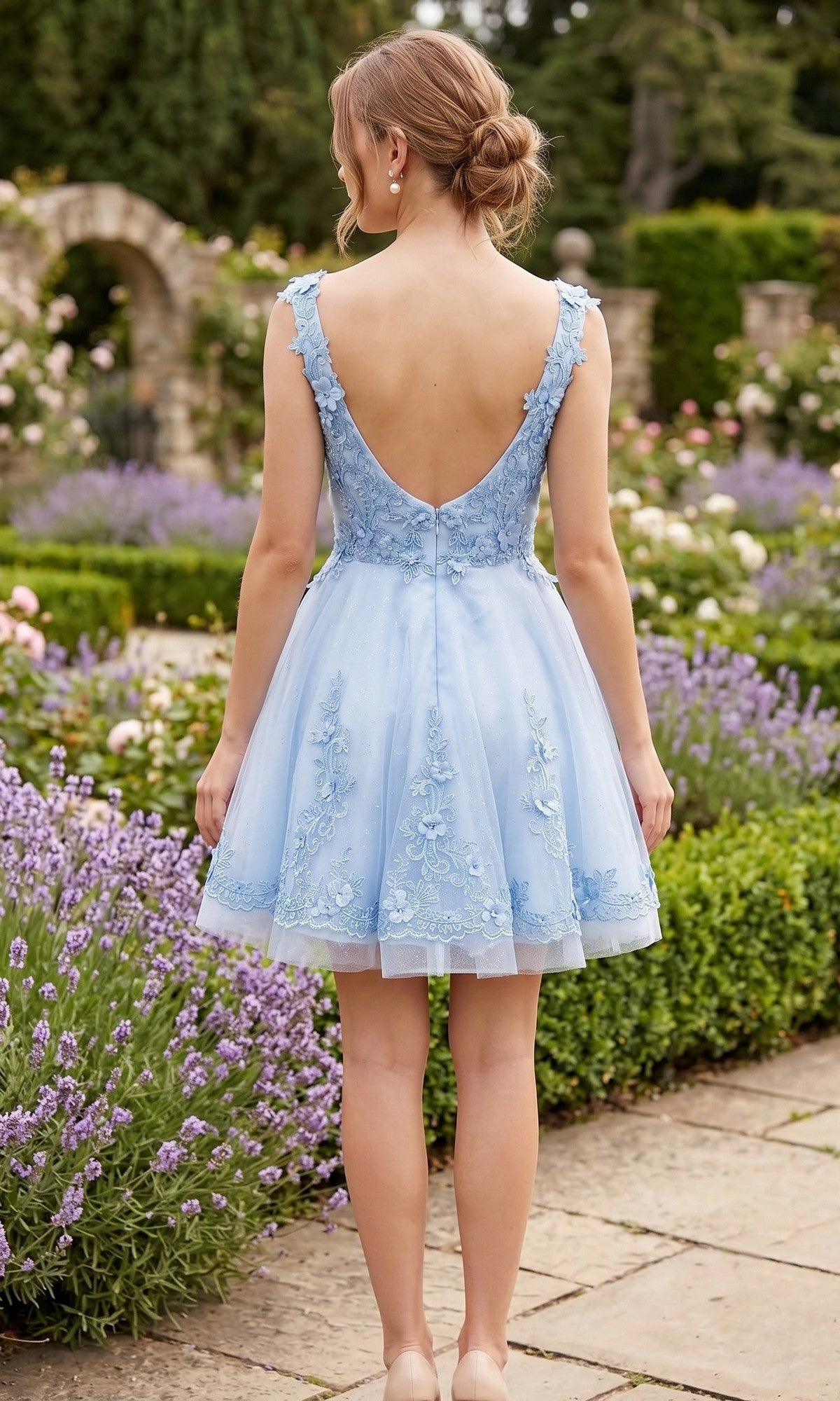 A woman with blonde hair in a low bun wears the Chicas C311 short homecoming dress in pastel blue with floral embroidery, standing on a garden path facing away from the camera.